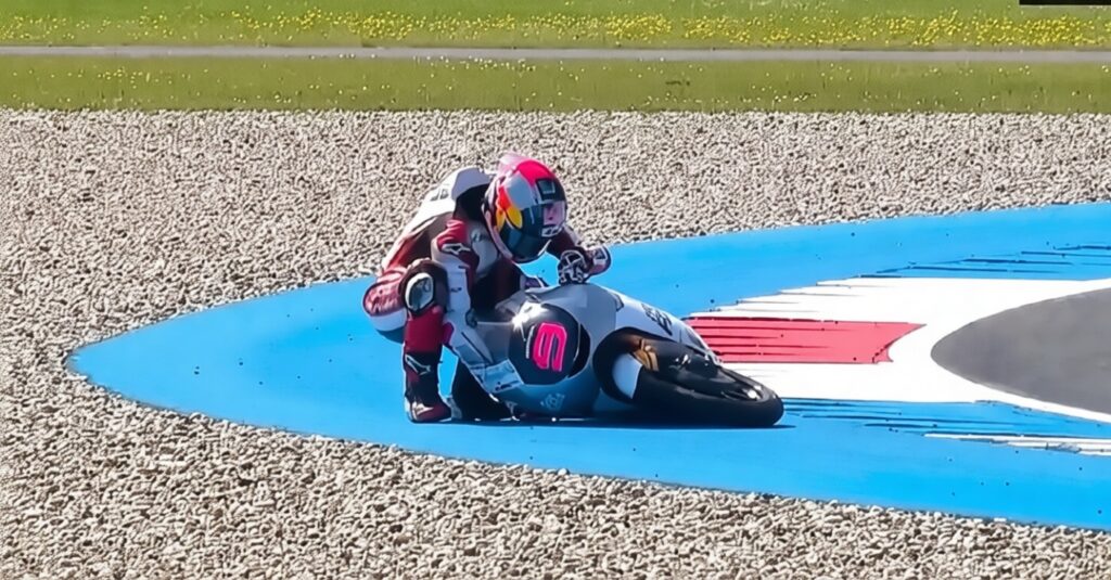 Moto racer in a pink helmet leans into a sharp blue-curved race track on a gravel run-off area, full speed ahead, white and red markings nearby