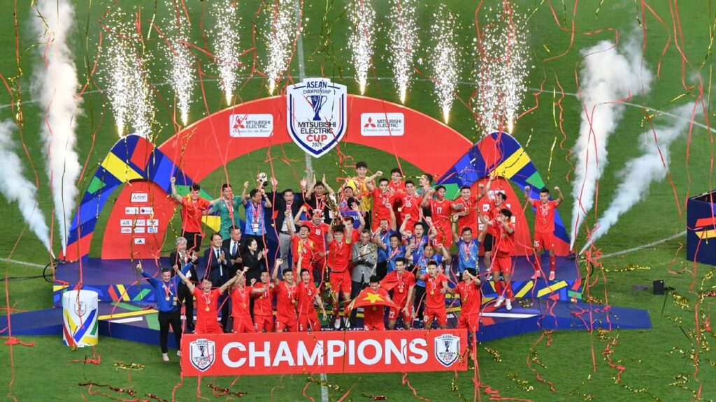 Vietnam lift AFF 2024 trophy - Mabur.co Red-clad team celebrates on a podium with trophy, confetti and fireworks overhead, and a large 'CHAMPIONS' banner in front.