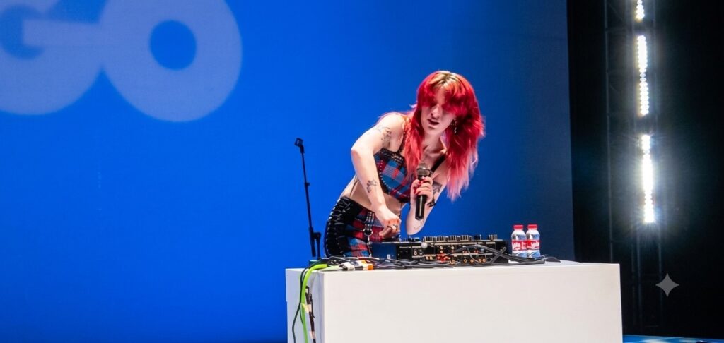 DJ with bright red hair performs on a blue stage, adjusting a mixer while holding a microphone at a white booth with cables and water bottles nearby