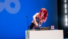 DJ with bright red hair performs on a blue stage, adjusting a mixer while holding a microphone at a white booth with cables and water bottles nearby