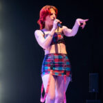 Female singer on stage singing into a microphone, wearing a colorful plaid outfit and pink suspenders, pointing to the audience.