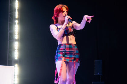 Female singer on stage singing into a microphone, wearing a colorful plaid outfit and pink suspenders, pointing to the audience.