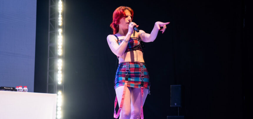 Female singer on stage singing into a microphone, wearing a colorful plaid outfit and pink suspenders, pointing to the audience.