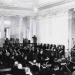 Black-and-white photo of a formal parliamentary session in a columned hall, delegates seated and a speaker at a podium.