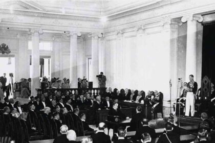 Black-and-white photo of a formal parliamentary session in a columned hall, delegates seated and a speaker at a podium.