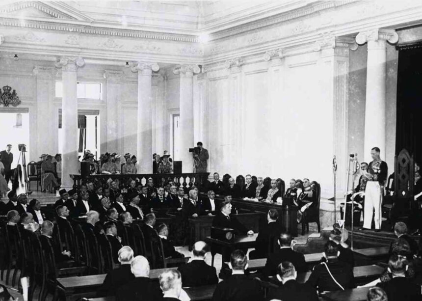Volksraad_04 - Mabur.co Black-and-white photo of a formal parliamentary session in a columned hall, delegates seated and a speaker at a podium.