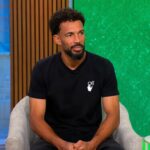 Man with short curly hair and beard sits in a light gray armchair in a TV studio, wearing a black t-shirt with a small white logo, green backdrop to the right and blue wall to the left.