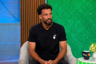 Man with short curly hair and beard sits in a light gray armchair in a TV studio, wearing a black t-shirt with a small white logo, green backdrop to the right and blue wall to the left.