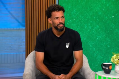 Man with short curly hair and beard sits in a light gray armchair in a TV studio, wearing a black t-shirt with a small white logo, green backdrop to the right and blue wall to the left.