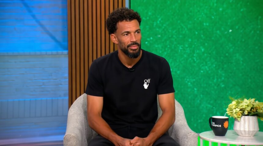 Man with short curly hair and beard sits in a light gray armchair in a TV studio, wearing a black t-shirt with a small white logo, green backdrop to the right and blue wall to the left.
