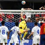 Soccer goalkeeper in yellow leaps to save ball at the goal as white-team players watch in front of the net.