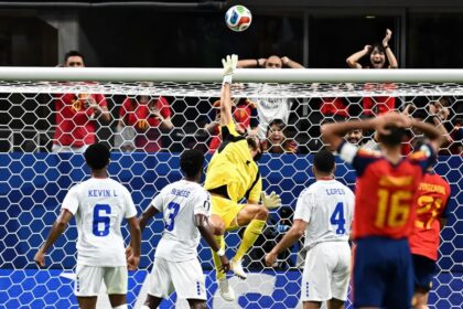Soccer goalkeeper in yellow leaps to save ball at the goal as white-team players watch in front of the net.