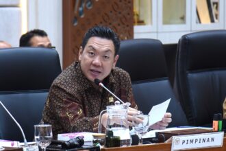 Man in a batik shirt speaks into a microphone at a conference table, with papers and glasses in front and a PIMPINAN nameplate nearby.