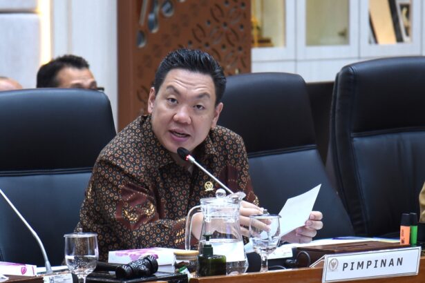 Man in a batik shirt speaks into a microphone at a conference table, with papers and glasses in front and a PIMPINAN nameplate nearby.