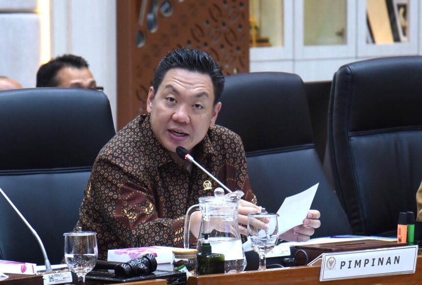 Man in a batik shirt speaks into a microphone at a conference table, with papers and glasses in front and a PIMPINAN nameplate nearby.