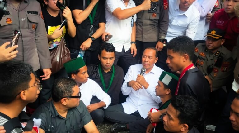 Group of men in uniforms and casual clothes seated in a circle as a man in a white shirt speaks into a microphone.