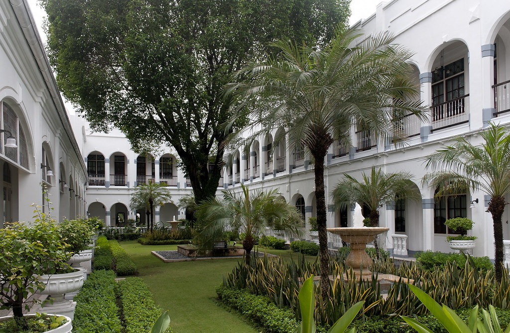 WhatsApp-Image-2024-03-01-at-153816-1-1297974420 - Mabur.co White, two‑story building courtyard with arches, a central fountain, and tropical plants.