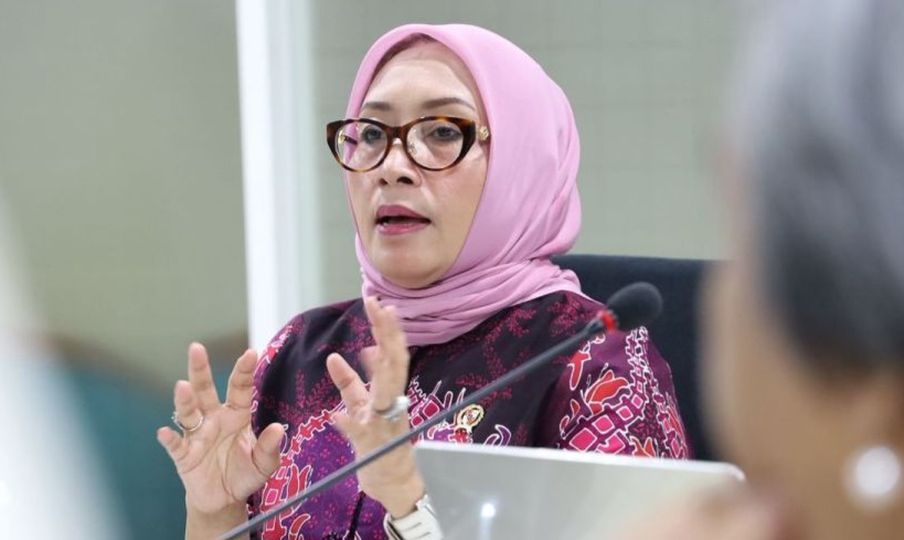 Woman in a pink hijab and glasses speaks at a meeting, gesturing with her hands.