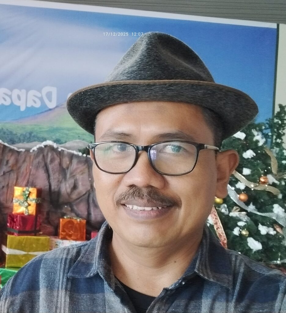 Smiling man wearing a gray fedora and glasses, standing in front of a Christmas tree with wrapped gifts in the background.