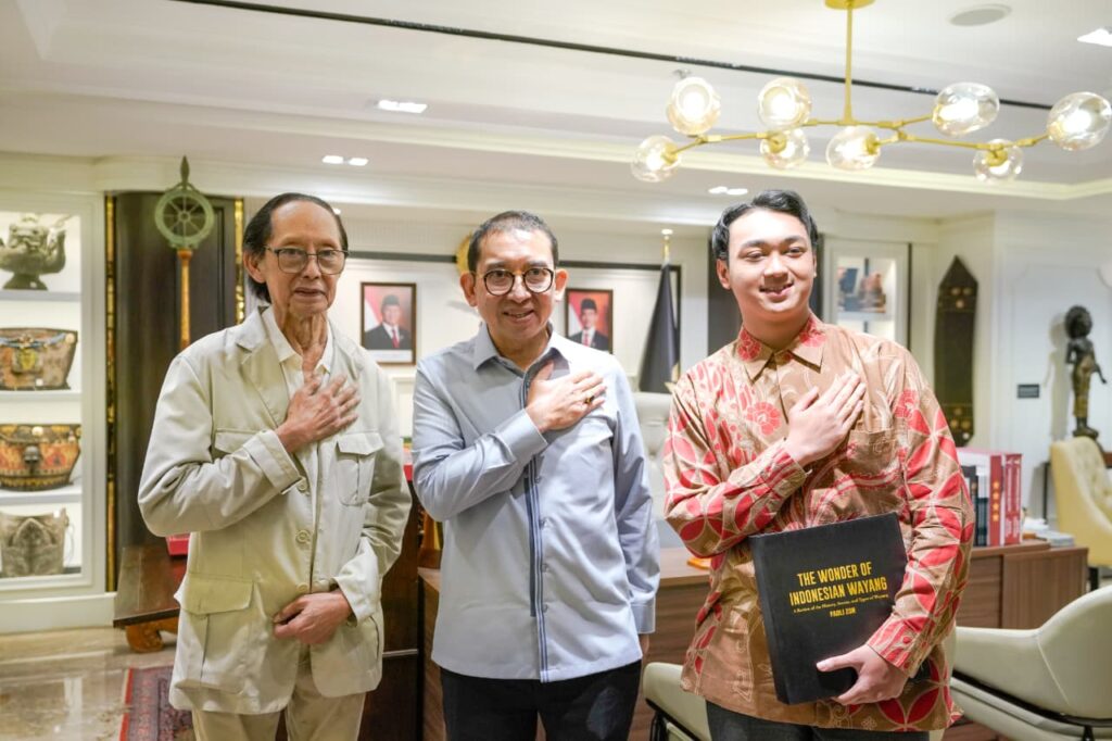 WhatsApp Image 2026-04-16 at 205532 - Mabur.co Three men stand side by side with their right hands over their chests in a formal room, the man on the right holding a book titled 'The Wonder of Indonesian Wayang'.
