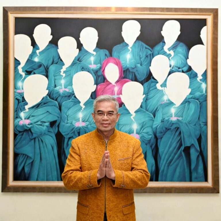 Man in an orange jacket stands with hands pressed together in front of a large painting featuring blue-robed, faceless figures holding candles (pink-hooded figure center).