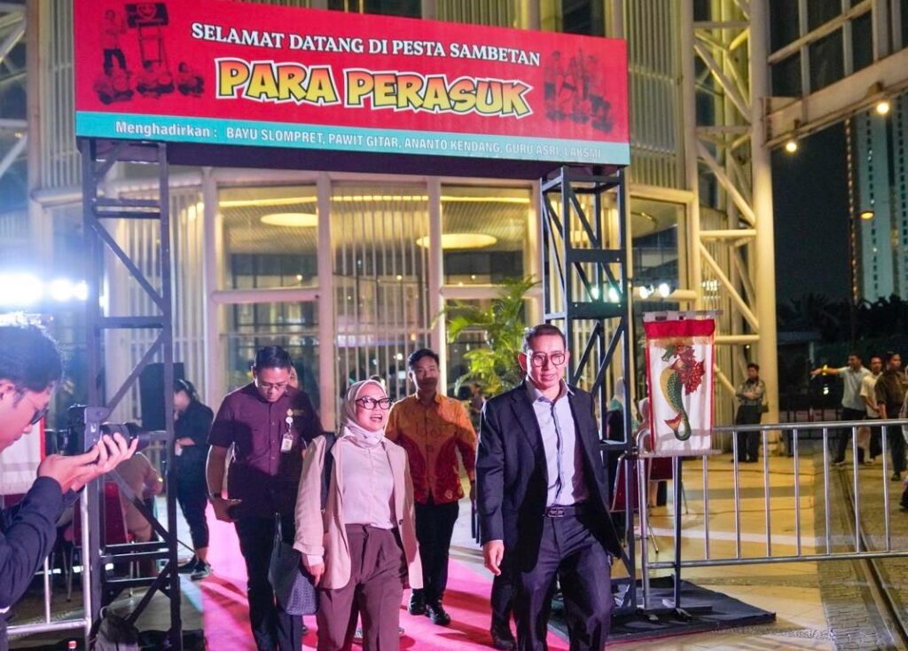 Group of people walking on a red carpet under a bright red banner that reads 'Para Perasuk' outside a building; photographer in the foreground captures the moment.