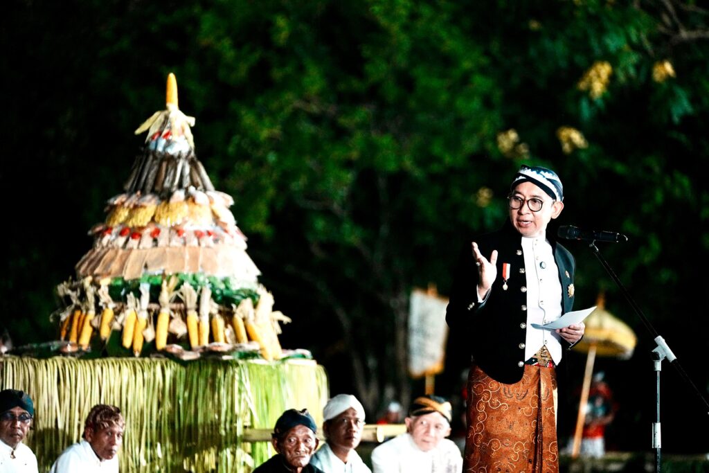 Man in traditional clothing speaks into a microphone during a night ceremony, with a tall corn-and-offering tower behind him.