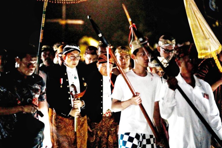 Night-time procession with young men in ceremonial attire holding staffs and banners.