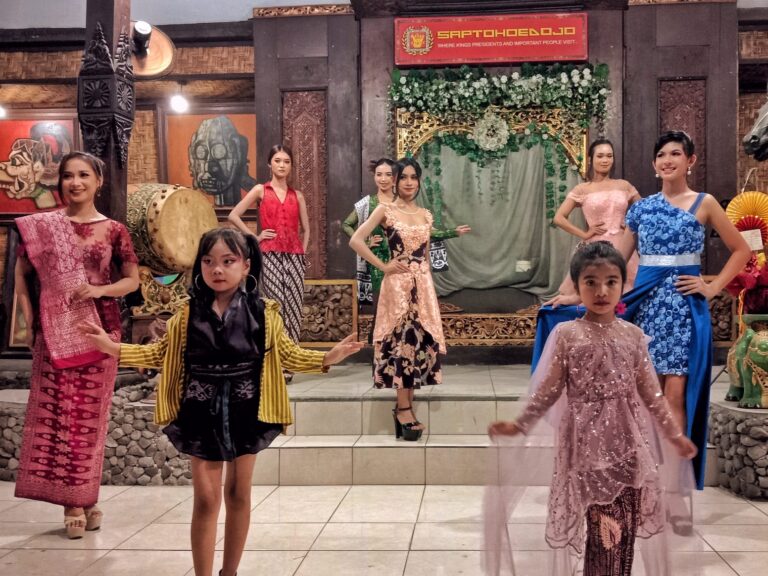 Group of young girls in colorful traditional dresses posing on a stage with ornate backdrop and sign overhead, likely at a cultural event.
