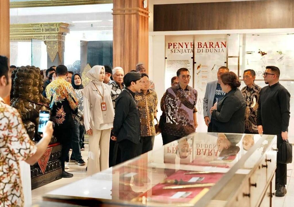 Group of people in batik and formal clothing stand in a museum hall, gathered around an exhibit case in the foreground