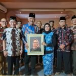 Group of people in batik attire posing together; the central man and woman hold a framed portrait of a woman wearing a hijab.