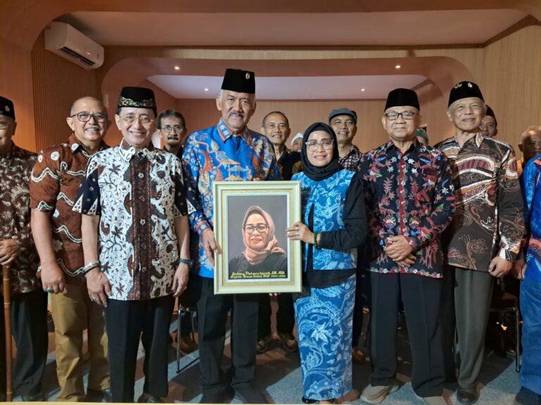 Group of people in batik attire posing together; the central man and woman hold a framed portrait of a woman wearing a hijab.