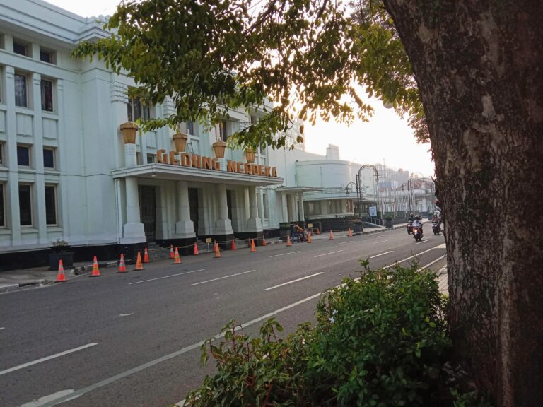 Illustration of a white building labeled 'Gedung Merdeka' beside a tree-lined street with orange traffic cones along the curb.