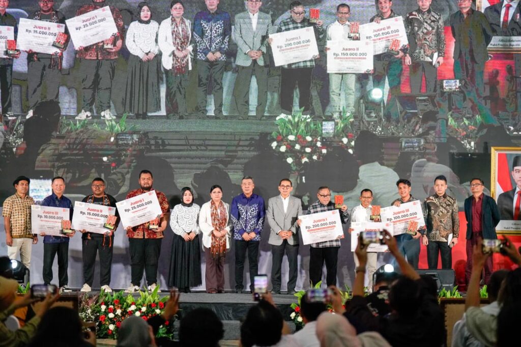 Group of award recipients on stage holding oversized prize checks during a ceremony, while audience members photograph the scene.
