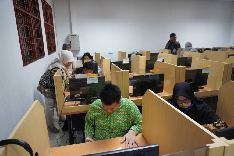 Computer lab with individual cubicles; people work at desktop computers, some wearing hijabs. A woman leans over a workstation in the foreground.