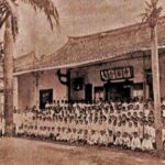 Small group photo of students in uniforms posing in front of a long traditional building with palm trees nearby.