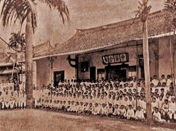 Small group photo of students in uniforms posing in front of a long traditional building with palm trees nearby.