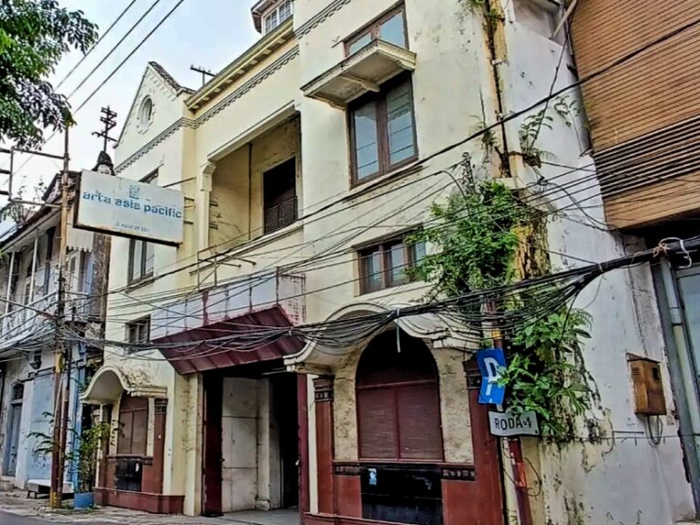 Two-story pale storefront building with arched entryways and a tangle of overhead wires along a narrow urban street, sign visible on the left reading 'arta asia pacific'.