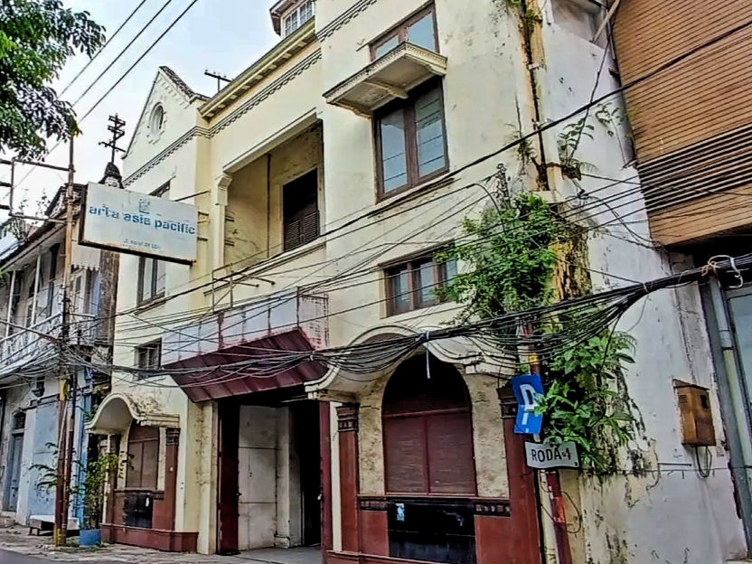 Two-story pale storefront building with arched entryways and a tangle of overhead wires along a narrow urban street, sign visible on the left reading 'arta asia pacific'.