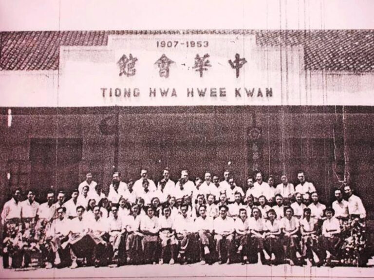Group photo of dozens of people in white shirts posing in front of a building with a sign displaying '1907-1953' and Chinese characters; appears to be a historical organization gathering.