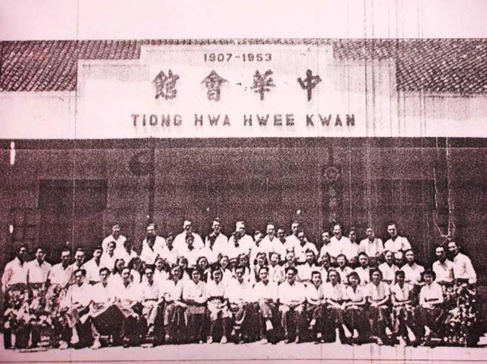 Group photo of dozens of people in white shirts posing in front of a building with a sign displaying '1907-1953' and Chinese characters; appears to be a historical organization gathering.