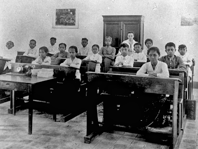 Black-and-white photo of young students seated at wooden desks in a classroom, with a teacher standing at the back near a cabinet.