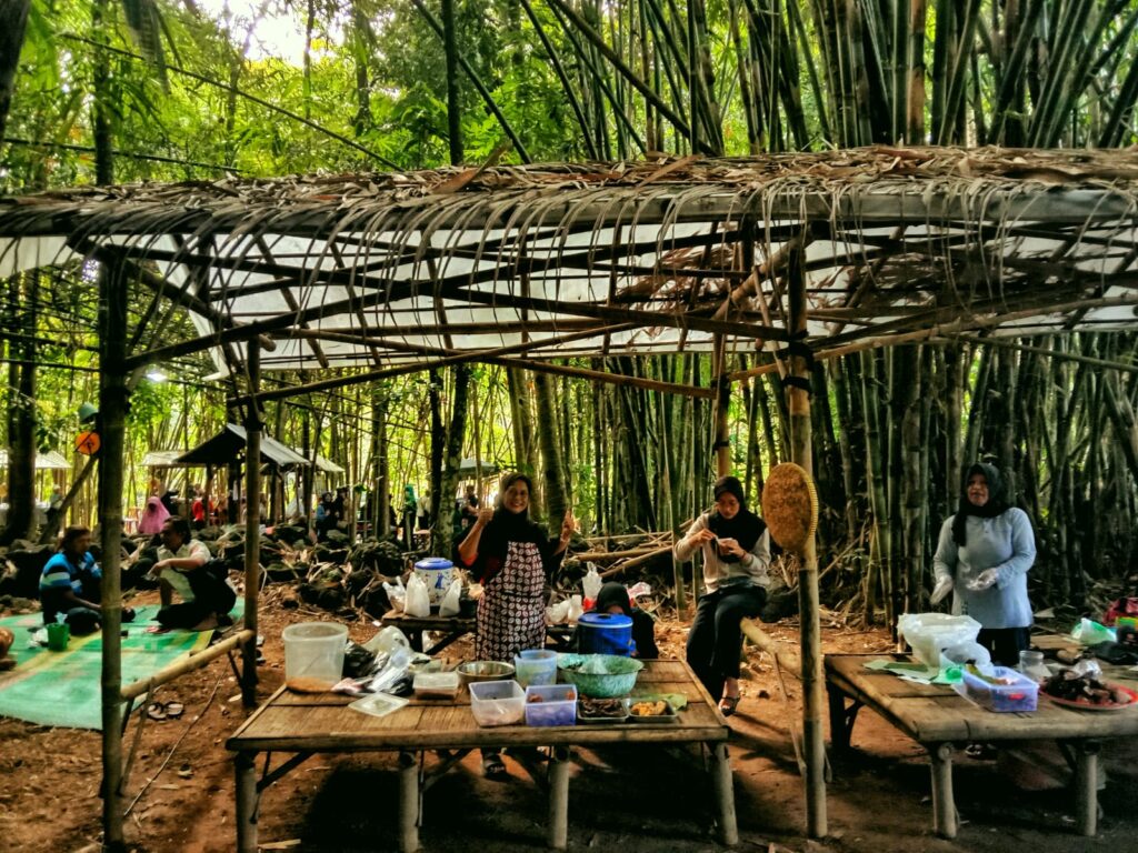 Group of people cooking and eating under a bamboo shelter with tables, baskets, and food containers in a forest setting