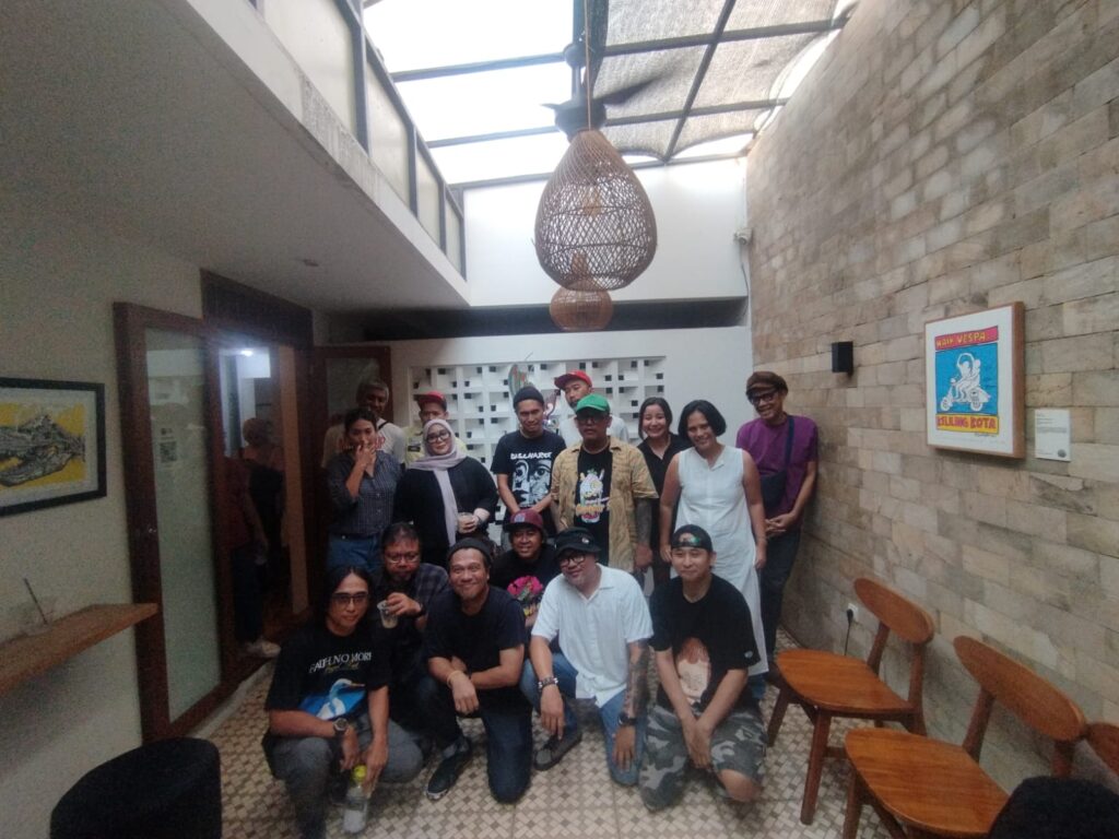 A group of about 15 people posing together in a narrow hallway with a large woven pendant light hanging from the ceiling and a stone wall on the right side.