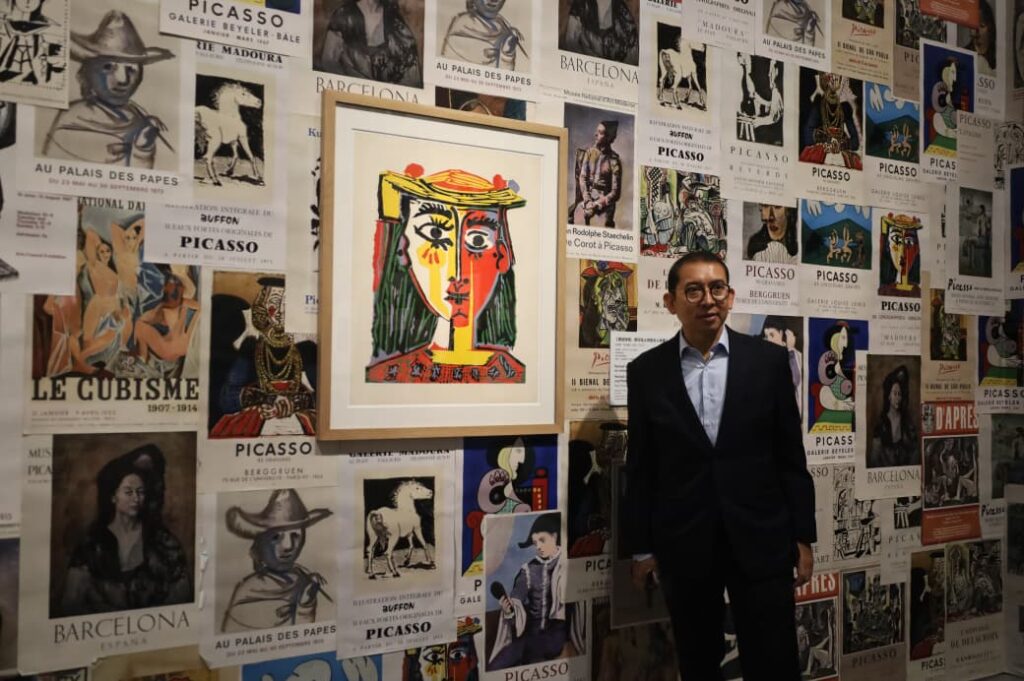 Man in a dark suit posing beside a framed colorful abstract portrait, with a wall covered in Picasso posters in the background.