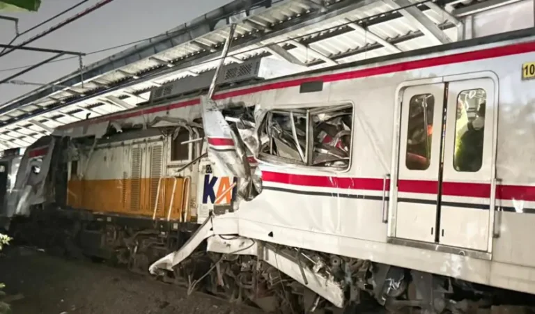 Damaged passenger train with crushed front end and shattered windows after a collision under a station canopy.