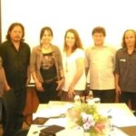 Eight adults stand side by side behind a conference table with papers and a floral centerpiece, posing for a group photo in a meeting room.