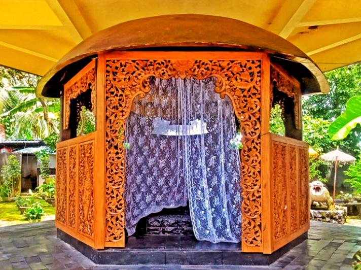 Ornate wooden pavilion with open carved doors and a waterfall feature inside, set in a tropical garden.