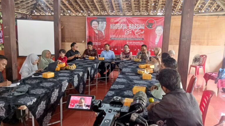 People sit around a U-shaped table in a hall; a red banner reading 'Ngobrol Bareng' dominates the background as a camera records the scene.