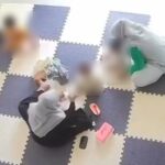 Overhead view of a room with checkered foam floor tiles; four blurred people sit on the floor in a circle, with a pink lunchbox and a bottle nearby.